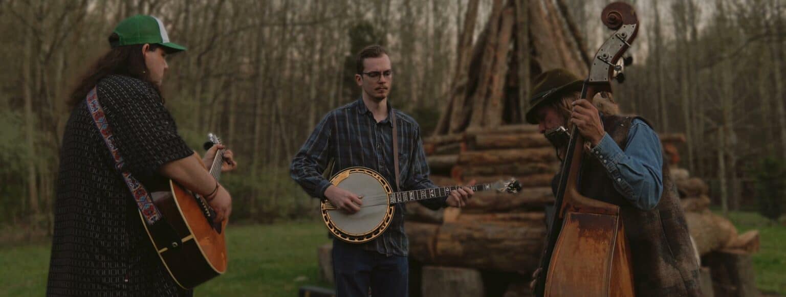 Musicians play banjo and guitar at an outdoor jam session by a log structure.