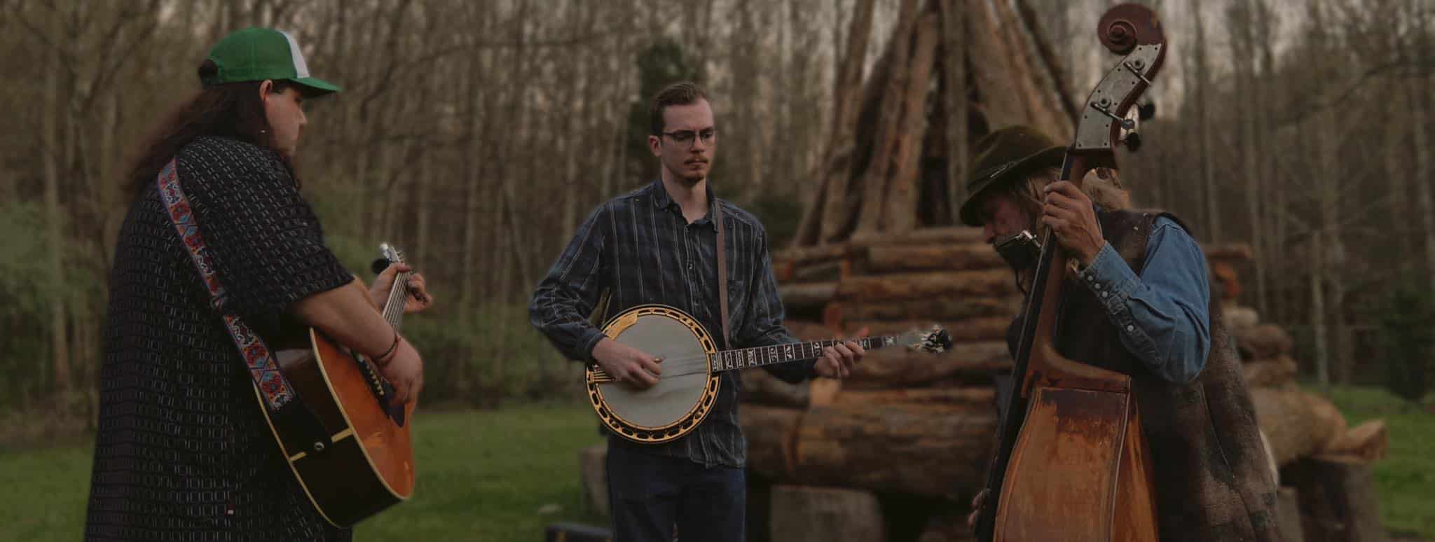 Musicians play banjo and guitar at an outdoor jam session by a log structure.