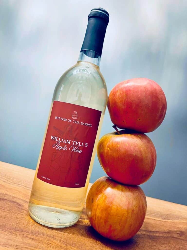 William Tells apple wine bottle next to stacked fresh apples on wooden surface.