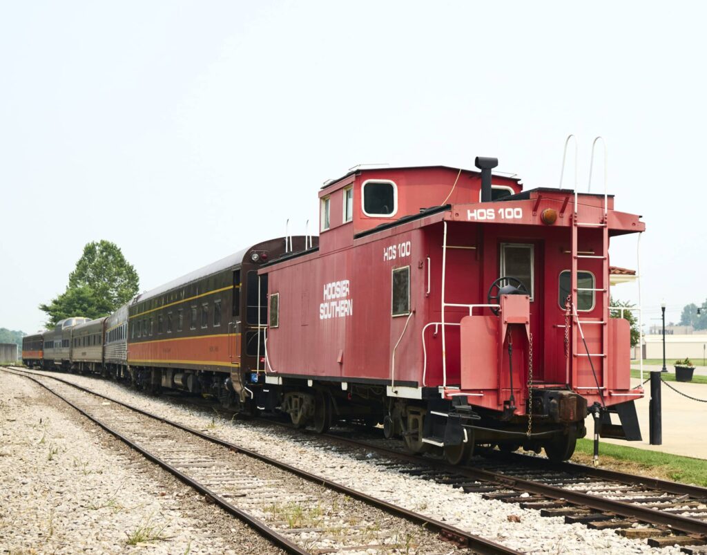 Red Hoosier Southern caboose HOS 100 parked beside vintage train cars on the tracks.