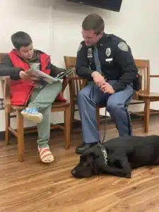 Child reads with a police officer and his dog, promoting community engagement and literacy.