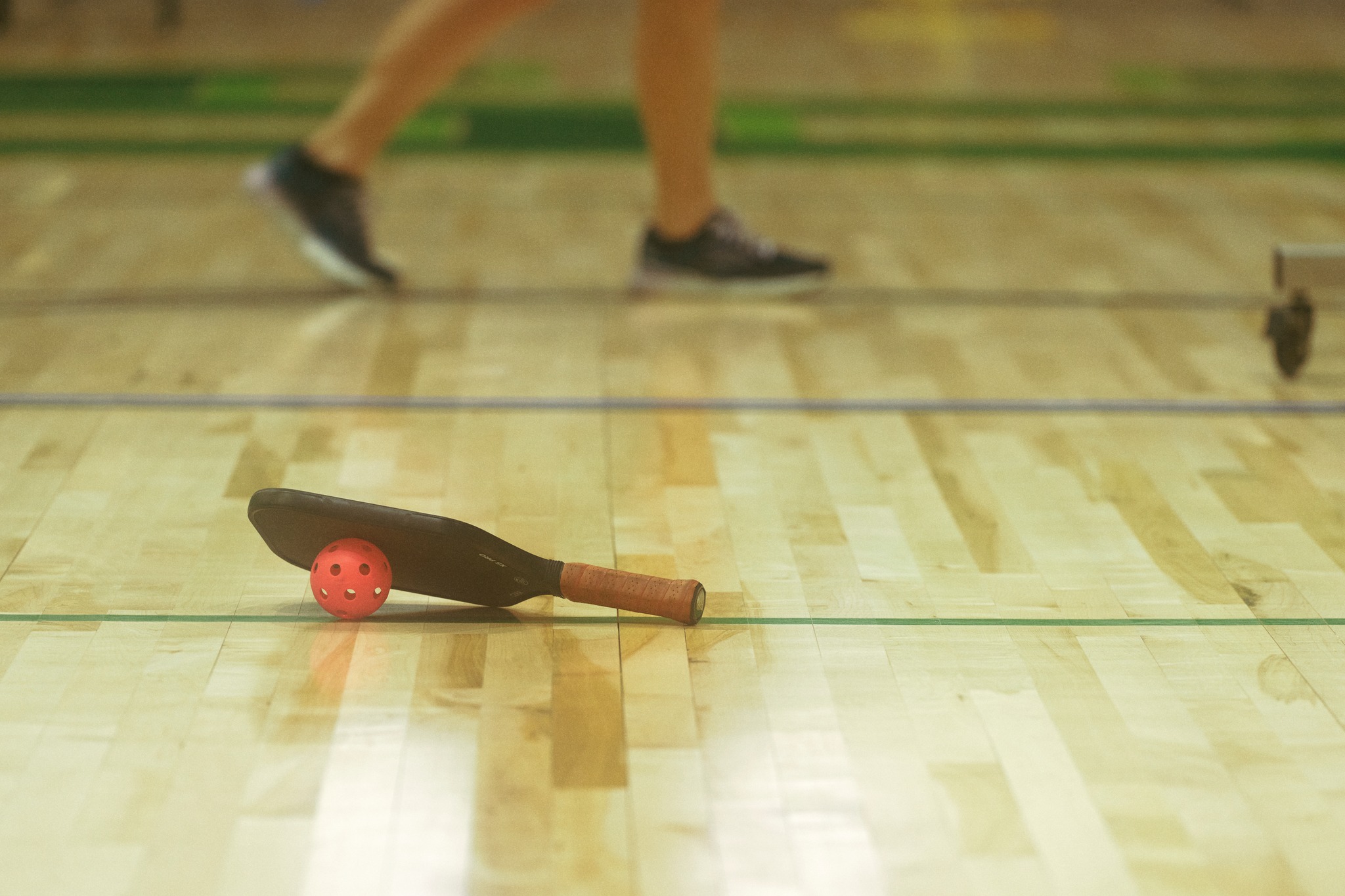 Pickleball paddle and ball resting on a wooden court floor.